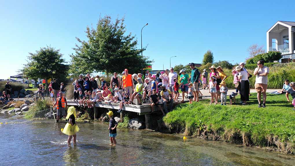 a crowd of people wait on the Silver Stream jetty for the yellow plastic ducks to arrive at the finish line