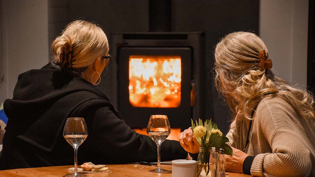 two woman enjoying a glass of wine in front of a warm wood burner