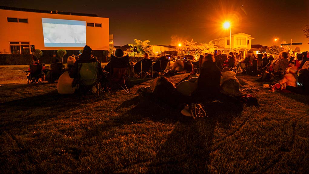 a large crowd of people gathered on a lawn at night time watching an outdoor movie